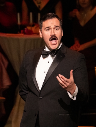 Student with mustache wearing a tuxedo singing opera on the stage with full cafe tables behind him