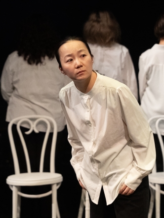 A student bending over a little while behind her are four white chairs and actors with their backs to her