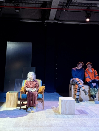 Five performers on stage surrounded by plywood boxes. Two are standing, two sit beside one another on a pair of boxes, and one woman sits in a chair with both hands on her knees.