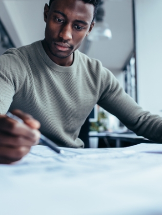 A man in a tan sweater sits at a desk in a bright office, focused on reviewing and marking documents with a pen.