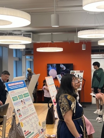 Students stand before research posters at tables in a bustling room. 