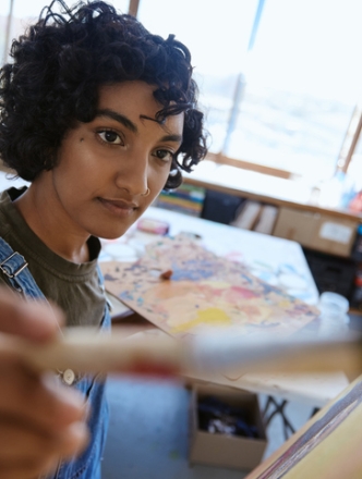 A young artist with curly hair wearing denim overalls focuses on a canvas, holding a paintbrush in the foreground with a wooden palette and sunlit studio in the background.