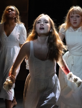 Three female students in white dresses on stage singing a dramatic song