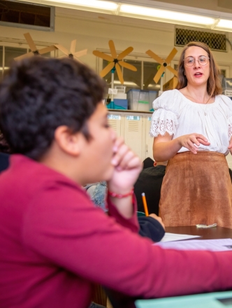 A teacher in the center of the classroom addresses a student holding a handout. 